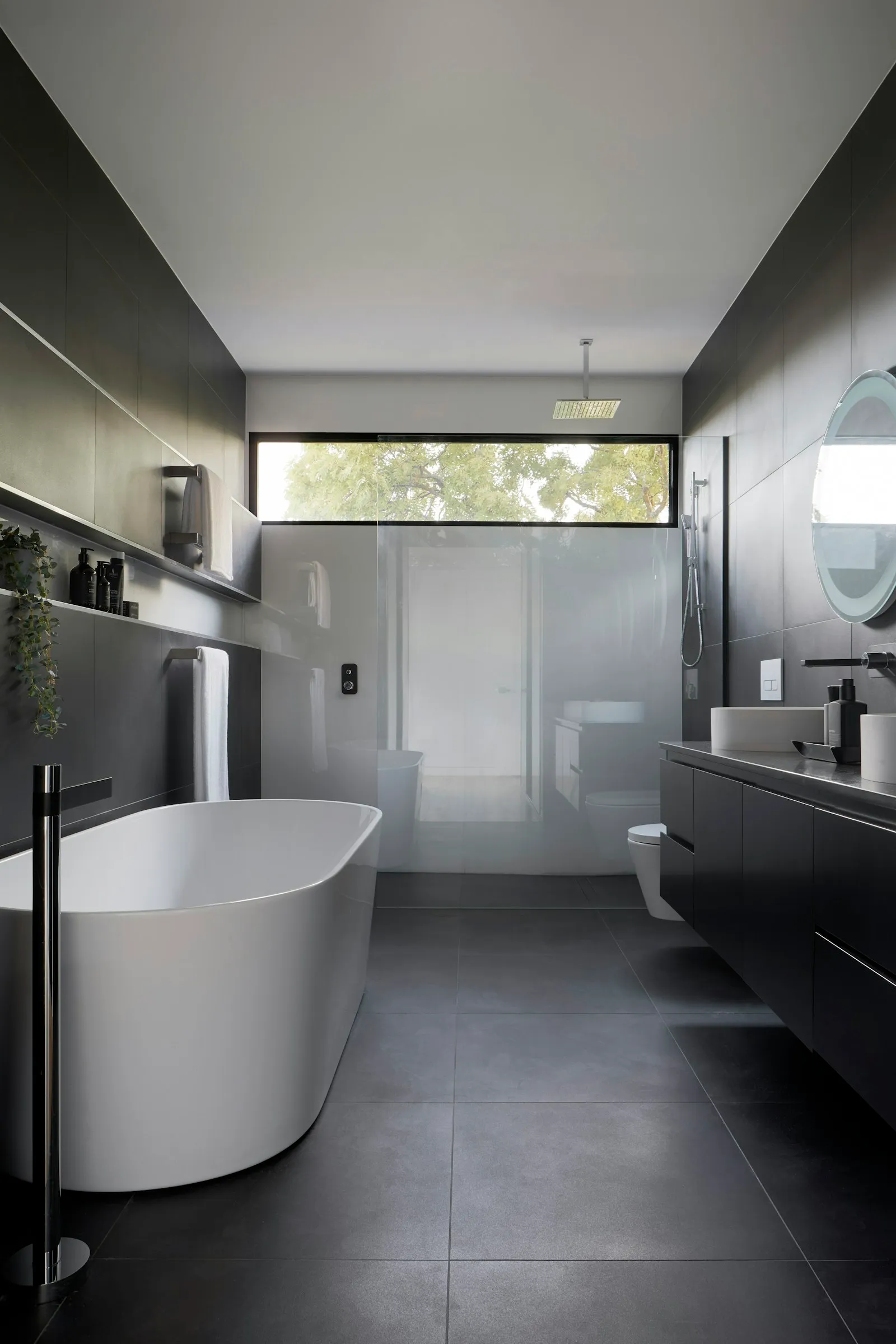 Kamar mandi modern gelap dengan bathtub freestanding putih dan pencahayaan tersembunyi